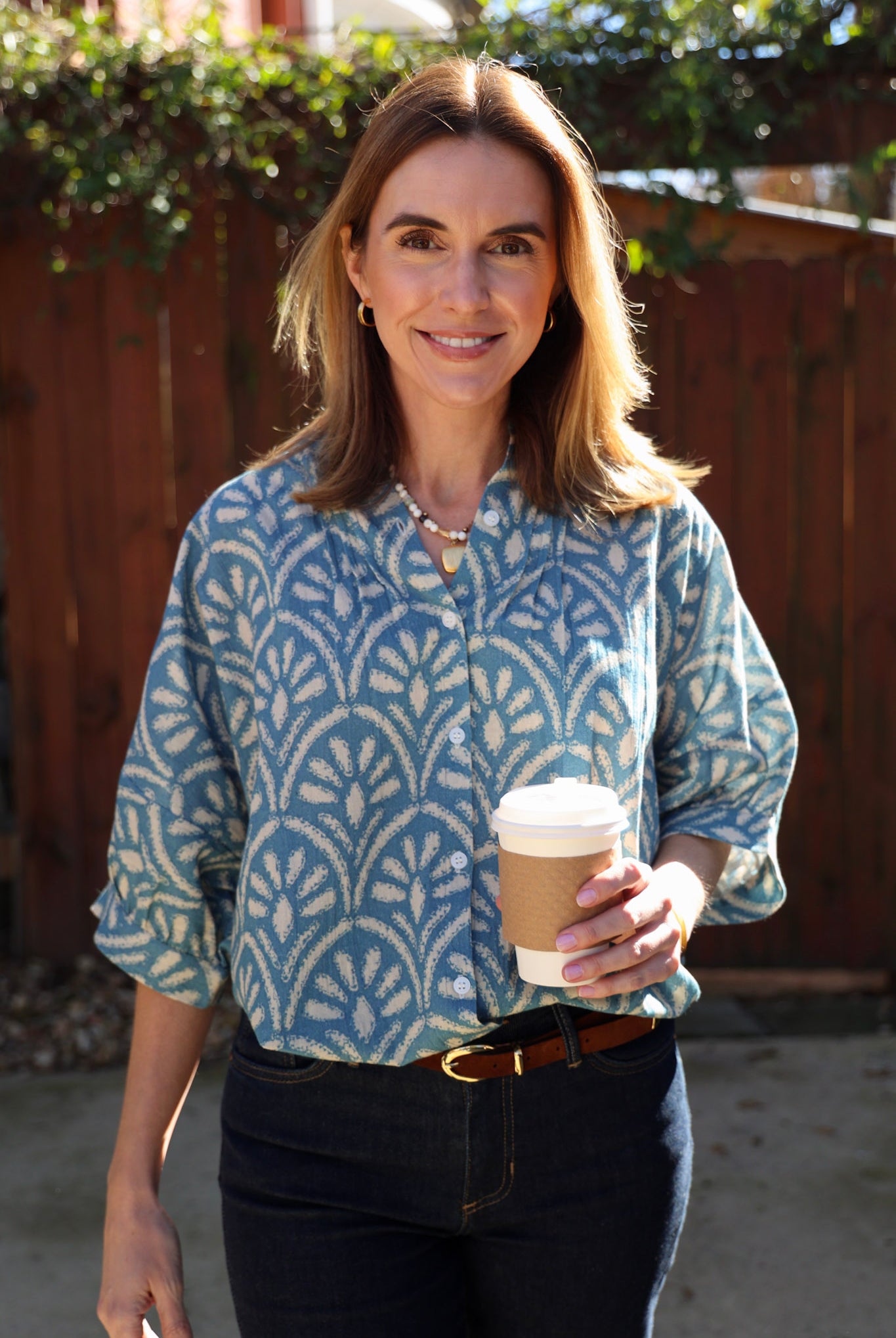 Woman holding a coffee cup outdoors with a wooden fence and greenery in the background wearing a blue blouse and jeans