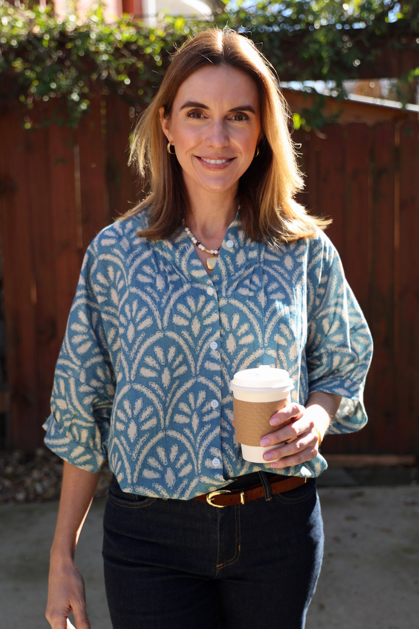 Woman holding a coffee cup outdoors with a wooden fence and greenery in the background wearing a blue blouse and jeans