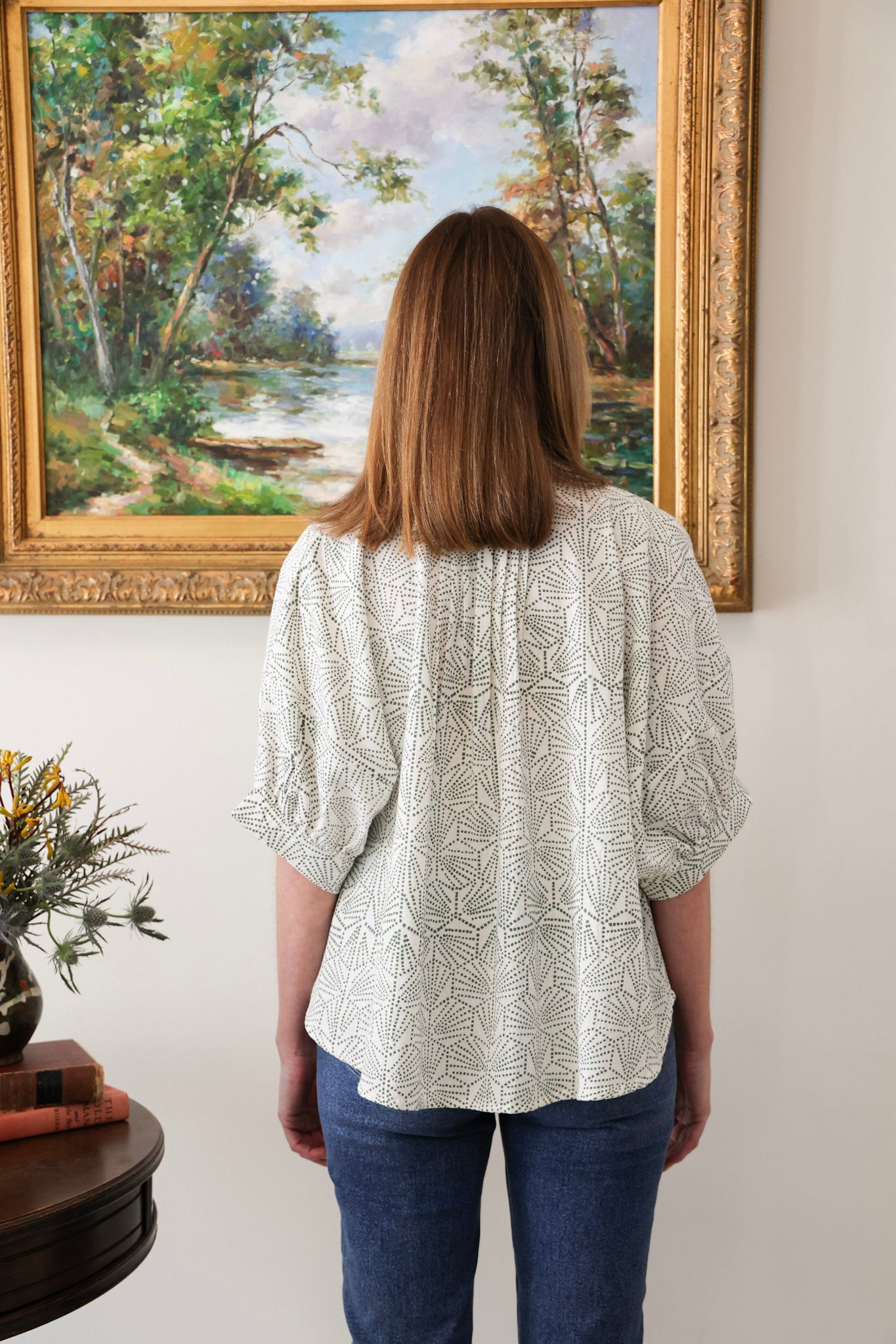 Woman wearing a patterned blouse standing in front of a large landscape painting.