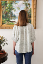 Woman wearing a patterned blouse standing in front of a large landscape painting.